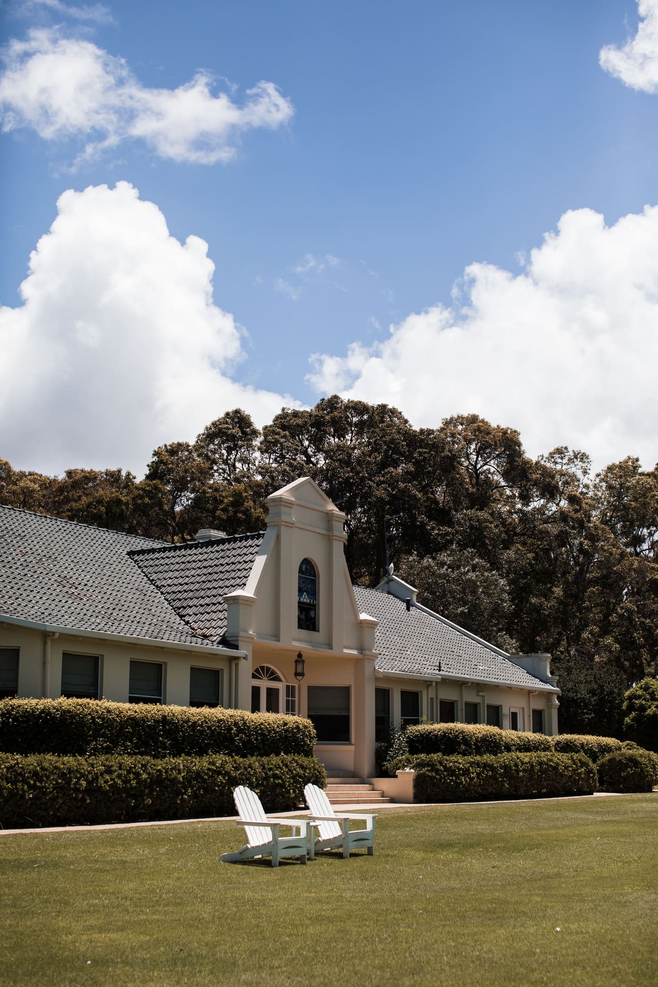 Modern home with white cupola, gray tile roof, manicured hedges, and Adirondack chairs on lawn