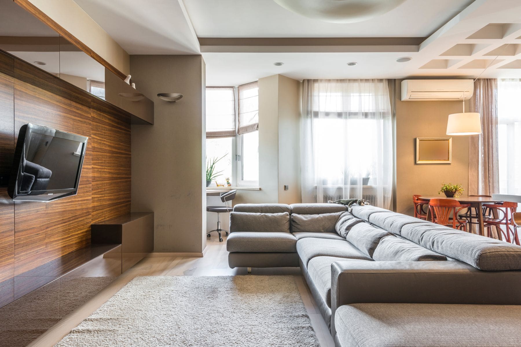 Modern living room with gray sectional sofa, wooden TV wall, large windows and open kitchen area