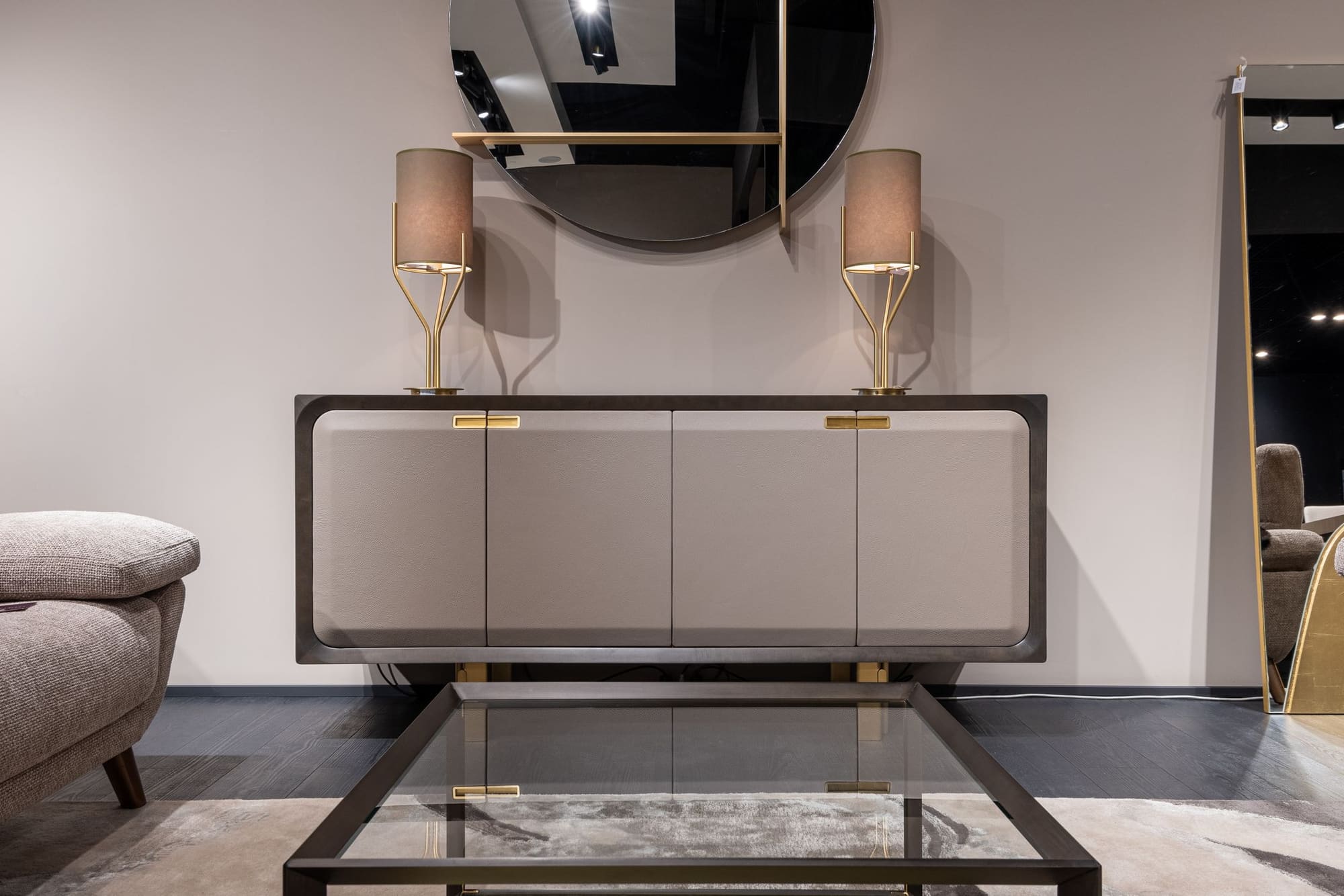 Modern bedroom furniture display with dark frame dresser, round mirror, brass lamps, and gray upholstered chair