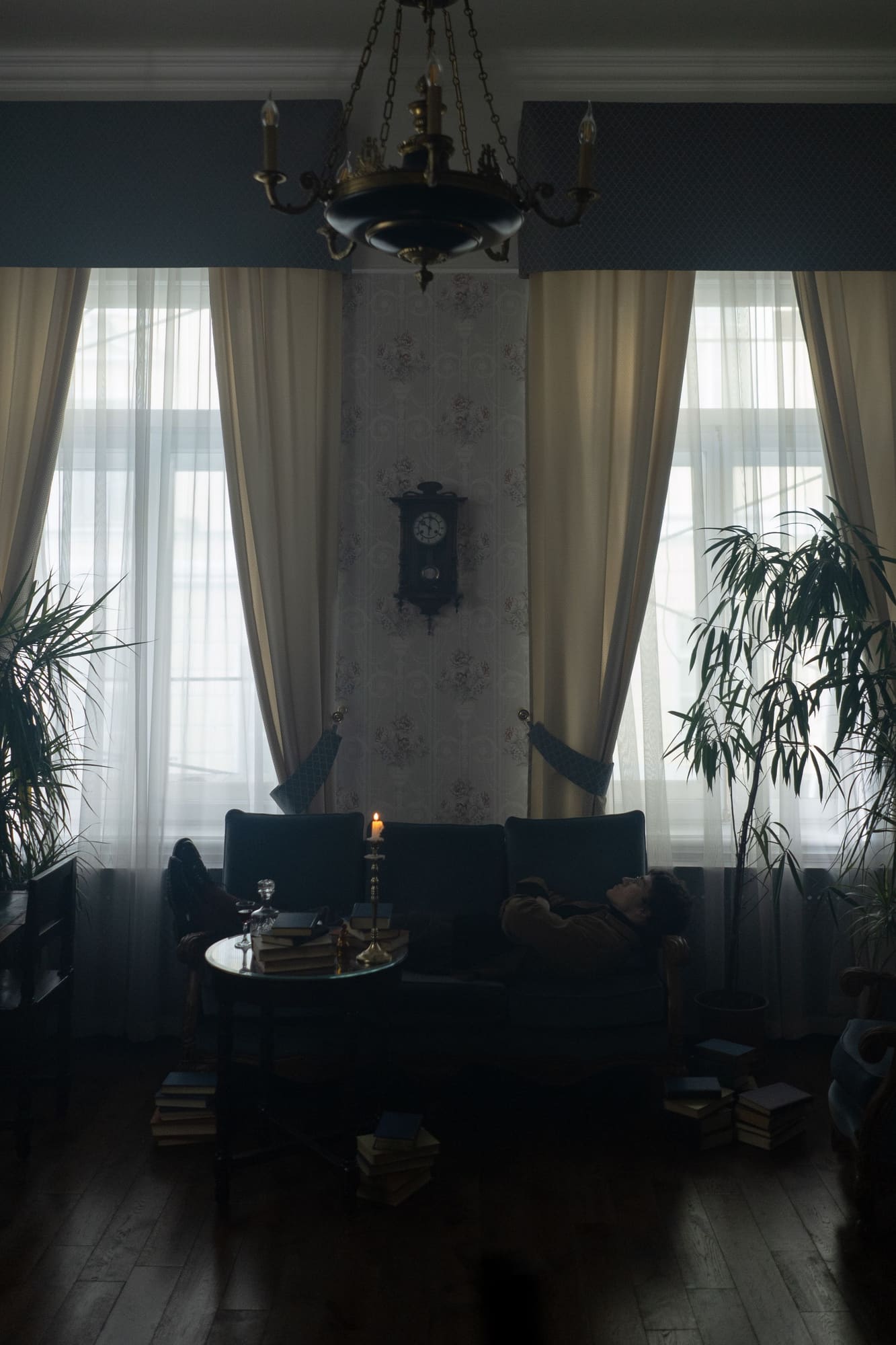 Elegant living room with black sofa, ornate chandelier, tall windows with cream curtains, and potted plants